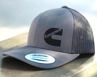 Può includere: Un cappello da camionista grigio e nero con un logo Cummins nero sul davanti. Il cappello ha una rete sul retro e una chiusura a scatto.
