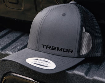 Puede incluir: Una gorra de camionero gris y negra con una parte trasera de malla negra. La parte delantera de la gorra tiene la palabra "TREMOR" bordada en negro.