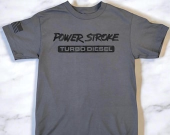 Peut inclure: Un t-shirt gris avec un motif noir qui dit "POWER STROKE TURBO DIESEL". Le motif est dans une police de caractères stylisée. Il y a un petit drapeau américain sur la manche gauche.