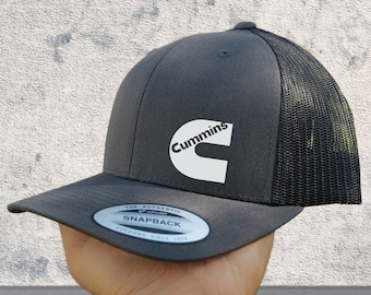 Può includere: Un cappello da camionista grigio e nero con un logo Cummins bianco e il testo "The Authentic Snapback Yupoong Since 1974".