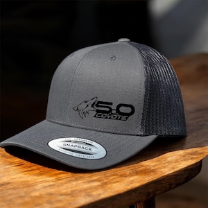 May include: Gray and black snapback trucker hat with the text "5.0 Coyote" and a coyote graphic. The hat has a mesh back and a curved brim. The hat has a snap closure and a Yupoong label.