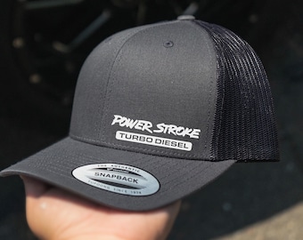 Peut inclure: Un chapeau de camionneur gris et noir avec un logo blanc qui dit "POWER STROKE TURBO DIESEL". Le chapeau a un dos en maille noire et un devant gris avec une étiquette blanche "The Authentic Snapback Yupoong Since 1974".