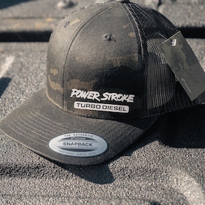 May include: A black and green camouflage snapback hat with the words "POWER STROKE TURBO DIESEL" in white. The hat has a mesh back and a curved brim. A tag is attached to the hat.
