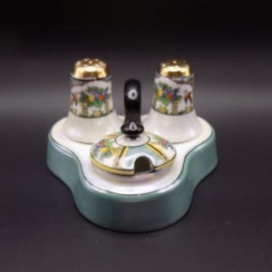 Puede incluir: Un juego de salero y pimentero de cerámica vintage con un tarro de condimentos a juego con tapa. El juego presenta detalles dorados y diseños florales sobre una base blanca, colocado sobre una bandeja verde claro. Los saleros y pimenteros tienen tapas doradas.