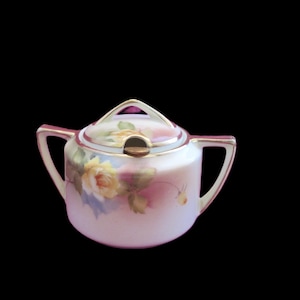 Puede incluir: Un azucarero de porcelana con tapa que presenta un diseño de rosa amarilla pintado a mano sobre un fondo rosa suave y blanco. El cuenco tiene dos asas laterales y un borde dorado, con una pequeña muesca en la tapa para colocar una cuchara.