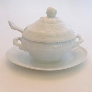 May include: White porcelain gravy boat with a lid and a small spoon. The boat has a decorative design and two handles. It is sitting on a matching saucer.