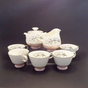 Op de afbeelding: Een vintage theeservies met een suikerpot met deksel, een melkkan en zes kopjes. De set is wit met een roze basis en rand, en een delicaat bloemmotief. De kopjes hebben een gouden rand.