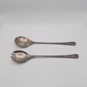 Puede incluir: Dos utensilios de servir de plata vintage. Uno es una cuchara grande y el otro es un tenedor con tres púas. Ambos tienen mangos largos y delgados con un diseño floral decorativo en el extremo.