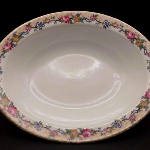 Op de afbeelding: Ovale, crèmekleurige serveerschaal met een bloemenrand. De rand heeft roze rozen, blauwe en gele accenten en groene bladeren op een beige achtergrond. De kom heeft een glanzende afwerking.