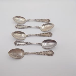 Könnte beinhalten: Fünf silberne Löffel mit verzierten, detaillierten Griffen liegen auf einer hellgrauen Oberfläche. Die Löffel haben ein klassisches, Vintage-Design mit polierter, reflektierender Oberfläche. Die Löffel sind wahrscheinlich zum Servieren oder Essen.