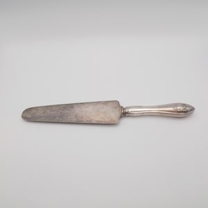 Puede incluir: Un cuchillo para pastel plateado vintage con una hoja redondeada y un mango adornado. El mango presenta un diseño floral decorativo. El cuchillo está diseñado para servir pasteles y repostería.