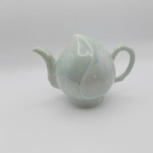Könnte beinhalten: Hellgrüne Keramik-Teekanne in Form einer Knospe, mit Ausguss, Henkel und Sockel. Die Oberfläche hat subtile blaue und weiße Akzente. Ein dekorativer Artikel für die Küche. Das Design der Teekanne erinnert an eine Blume.