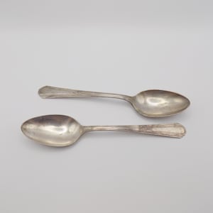 May include: Two antique silver spoons with oval bowls and decorative handles are displayed against a white background. The spoons are likely made of sterling silver and show signs of age and use. The spoons are a classic example of vintage silverware.