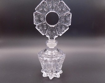 Può includere: Un flacone di profumo vintage trasparente con un tappo decorativo a motivo a stella. La bottiglia ha una base sfaccettata a forma di fiore e una parte superiore circolare. Il vetro è trasparente e il design è intricato, che mostra uno stile classico ed elegante.