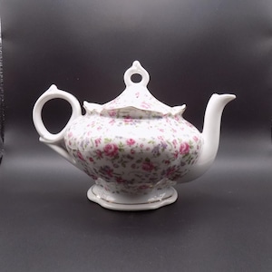May include: White porcelain teapot with a floral pattern of pink and purple roses. The teapot has a curved spout, a rounded body, and a loop handle. The lid has a small handle and gold trim.