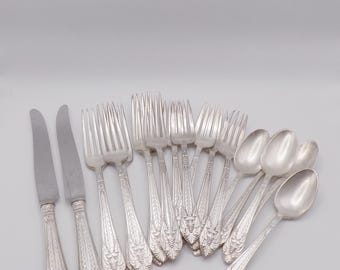 May include: A collection of silver-colored flatware, including knives, forks, and spoons, arranged on a white surface. The utensils have ornate floral designs on the handles. The set includes two knives, several forks, and multiple spoons.