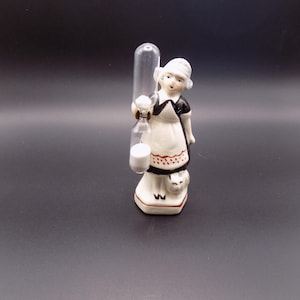 以下が含まれることがあります： 伝統的なオランダの衣装を着た女性のヴィンテージセラミックフィギュア。ガラス容器とミルクジャグを持っています。フィギュアは主に白で、黒と赤のアクセントがあります。フィギュアのベースには猫がいます。