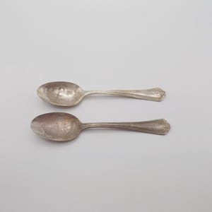 May include: Two antique silver spoons are shown on a light grey background. The spoons have an aged appearance, with oval bowls and decorative handles. The spoons are likely vintage silverware.
