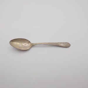 Può includere: Un cucchiaio d'argento con una coppa arrotondata e un manico decorativo. Il cucchiaio è realizzato in un materiale metallico lucido ed è posto su uno sfondo bianco. Il cucchiaio sembra essere vintage o antico.
