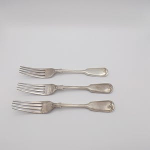 Puede incluir: Tres tenedores antiguos de plata, con púas y mangos en forma de corazón, están dispuestos horizontalmente sobre un fondo blanco. Los tenedores son de un metal brillante y reflectante, probablemente plata o una aleación plateada. El diseño es sencillo y elegante.