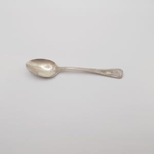 Puede incluir: Una cuchara de plata con un cuenco redondeado y un mango decorativo. La cuchara es de metal brillante y está sobre un fondo blanco liso. Un utensilio clásico para comer.