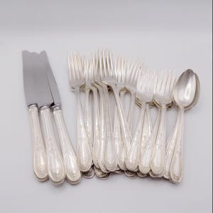 Puede incluir: Un conjunto de cubiertos plateados, que incluye cuchillos, tenedores y cucharas, dispuestos sobre una superficie blanca. Los cuchillos tienen una hoja redondeada y mangos ornamentados. Los tenedores y cucharas también tienen mangos decorativos. Los cubiertos parecen ser antiguos.