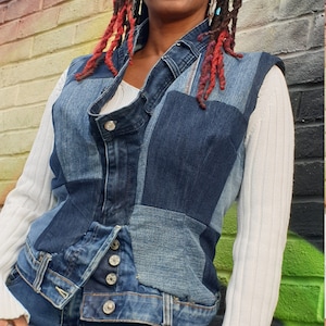 May include: A woman wearing a white long-sleeved shirt and a denim vest made of different shades of blue denim. The vest has a button closure and a cinched waist. The woman is wearing blue jeans and has red and black braided hair.