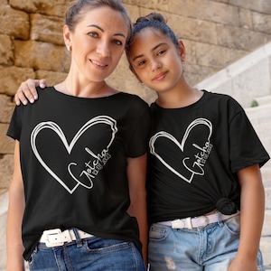 May include: Two black t-shirts with a white heart design and the word "Gotcha" with the date 12.08.2023. The shirts are worn with blue jeans and denim shorts. The background is a stone wall and steps.