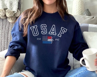 May include: Navy blue sweatshirt with the text "USAF" in white block letters, with a red, white, and blue graphic and the text "EST 1947". The sweatshirt is worn with ripped blue jeans. A white mug is held in the hand.