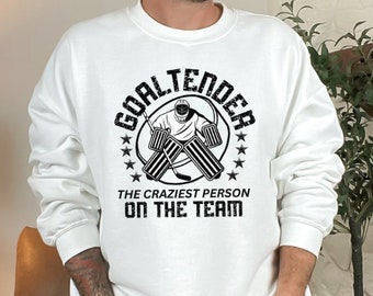 Op de afbeelding: Wit sweatshirt met een zwarte afbeelding van een hockeykeeper met de tekst "Goaltender The Craziest Person On The Team"