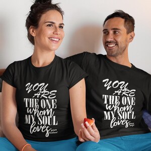 K&ouml;nnte beinhalten: Ein Paar tr&auml;gt passende schwarze T-Shirts mit wei&szlig;em Text, der "You are the one whom my soul loves" lautet. Sie sitzen auf einem Bett mit einer wei&szlig;en Bettdecke und blauen Laken.