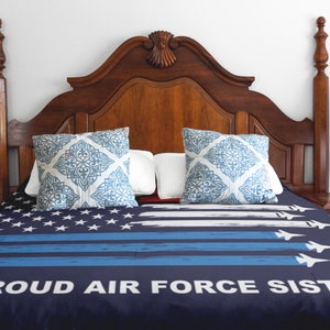 Op de afbeelding: Een marineblauwe en witte bedsprei met een verweerd Amerikaans vlagontwerp en de tekst "PROUD AIR FORCE SISTER". De bedsprei ligt op een bed met een houten hoofdeinde en twee blauwe en witte gepatroneerde kussens.