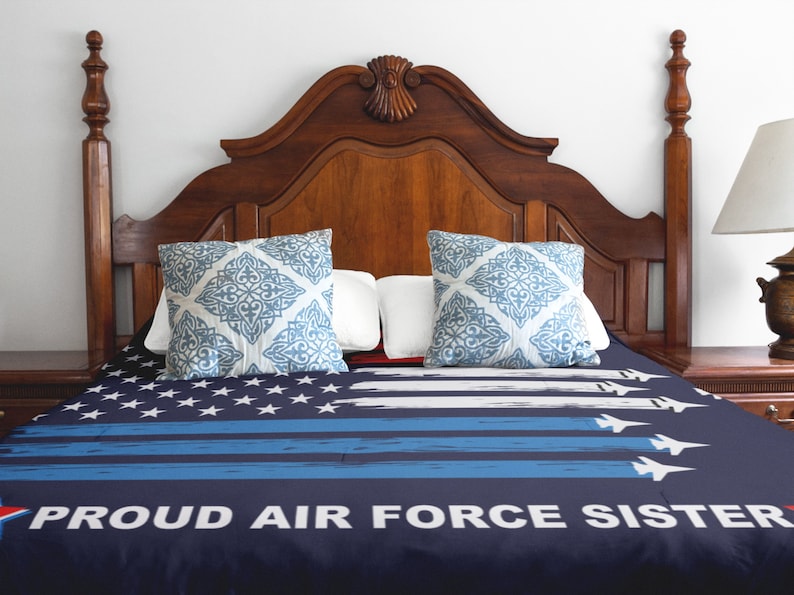 Op de afbeelding: Een marineblauwe en witte bedsprei met een verweerd Amerikaans vlagontwerp en de tekst "PROUD AIR FORCE SISTER". De bedsprei ligt op een bed met een houten hoofdeinde en twee blauwe en witte gepatroneerde kussens.