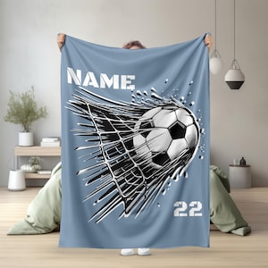 Könnte beinhalten: Hellblaue Decke mit einem schwarz-weißen Fußball-Design, das durch ein Netz bricht. Das Wort "NAME" und die Zahl "22" sind weiß. Die Decke wird von einer Person gehalten.