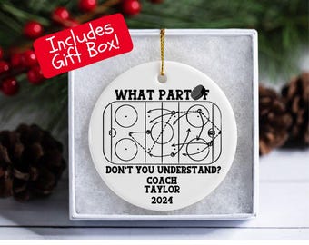 May include: A white ceramic hockey-themed ornament with a gold string for hanging. The ornament features a hockey rink diagram and the text "WHAT PART DON'T YOU UNDERSTAND? COACH TAYLOR 2024". Includes a gift box.