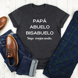 May include: A dark gray t-shirt with the words "PAPÁ, ABUELO, BISABUELO, Sigo mejorando" in white. The shirt is laid out with a pair of brown leather shoes, blue jeans, and a blue and green plaid button-down shirt.
