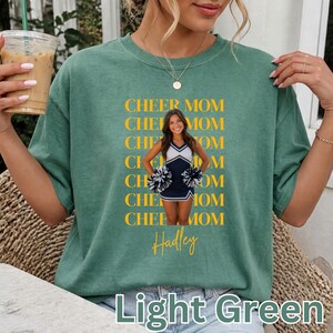 Könnte beinhalten: Ein salbeigrünes Kurzarm-T-Shirt mit einem sich wiederholenden gelben Textmuster, das CHEER MOM lautet. Die mittlere Grafik zeigt eine Cheerleaderin in einer marineblau-weißen Uniform mit Pompons, darunter steht der Name Hadley in Gelb.
