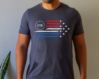 May include: Navy blue t-shirt featuring a patriotic design. The graphic includes a stylized American flag with red, white, and blue stripes, a circle of stars with the number "1776", and a formation of white jets.