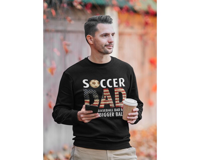 Soccer dad sweatshirt with American flag design and soccer ball graphic, funny soccer dad pullover worn outdoors in autumn setting