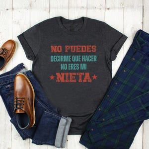 Könnte beinhalten: Dunkelgraues T-Shirt mit dem spanischen Text "NO PUEDES DECIRME QUE HACER NO ERES MI NIETA" in Rot und Türkis. Das Shirt wird mit blauen Jeans, einem karierten Hemd und braunen Lederschuhen präsentiert.