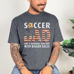 Funny soccer dad t shirt with American flag design and soccer ball graphic, men’s gray tee worn by model in studio setting