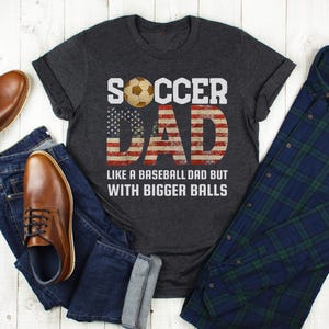 Funny soccer dad t shirt flat lay with jeans, boots, and flannel shirt, American flag soccer dad design on dark heather tee
