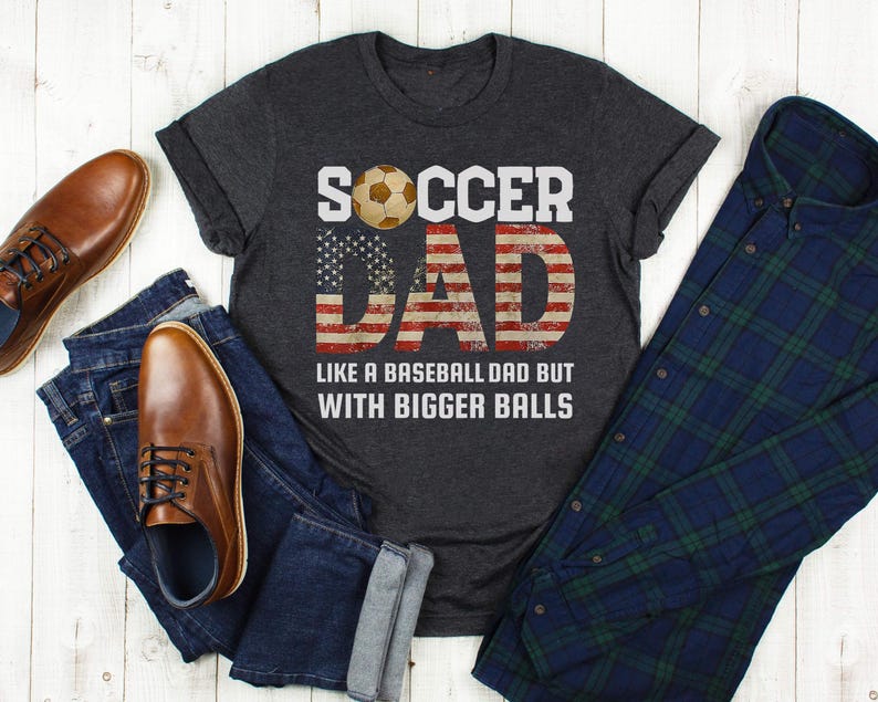Funny soccer dad t shirt flat lay with jeans, boots, and flannel shirt, American flag soccer dad design on dark heather tee