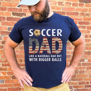 Funny soccer dad shirt with patriotic American flag lettering and soccer ball design, men’s navy t shirt worn outdoors against brick wall