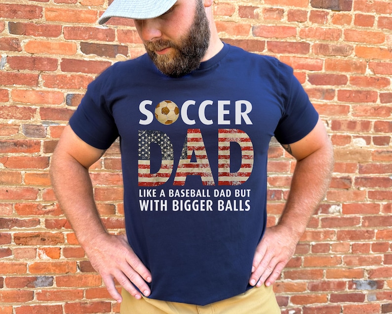Funny soccer dad shirt with patriotic American flag lettering and soccer ball design, men’s navy t shirt worn outdoors against brick wall