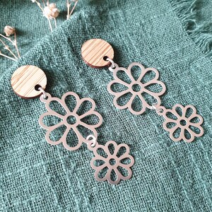 May include: A pair of silver-colored flower earrings with wooden circular tops. The earrings feature a large flower design connected to a smaller flower. The earrings are displayed on a teal-colored fabric.