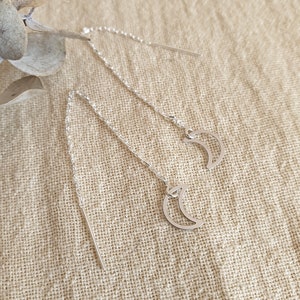 May include: Silver threader earrings with a crescent moon pendant. The earrings have a fine chain and a straight bar for insertion. The earrings are displayed on a neutral, textured surface, with a sprig of dried foliage in the background.