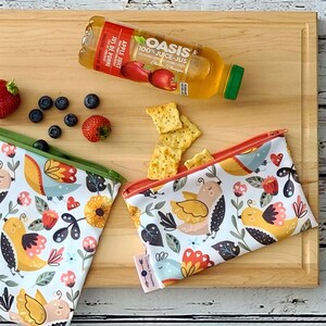 Peut inclure: Deux sacs à collation réutilisables avec un fond blanc et un motif coloré d'oiseaux et de fleurs. Les sacs ont une fermeture à glissière et sont faits de tissu imperméable. Les sacs sont sur une planche à découper en bois avec une bouteille de jus de pomme, des craquelins et des fruits.
