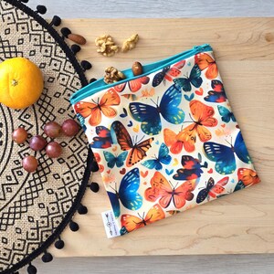 Peut inclure: Un sac à collation réutilisable blanc avec une fermeture éclair turquoise et un imprimé de papillons colorés. Le sac est posé sur une surface en bois avec un tapis à motifs noir et blanc et un bol de raisins en arrière-plan.