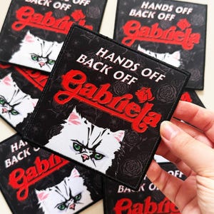 Puede incluir: Parche cuadrado negro con el texto "HANDS OFF BACK OFF Gabriela" en rojo, con un gato blanco de ojos verdes. El fondo es negro con patrones de rosas. Se ven varios parches.